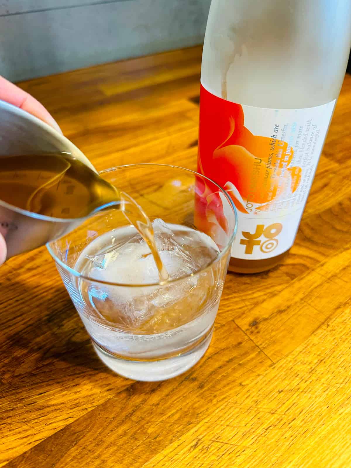 Pale peach colored liquid being poured from a steel measuring jigger into a rocks glass with a large ice cube next to a bottle of umeshu sake with a large red and orange flower on the label.