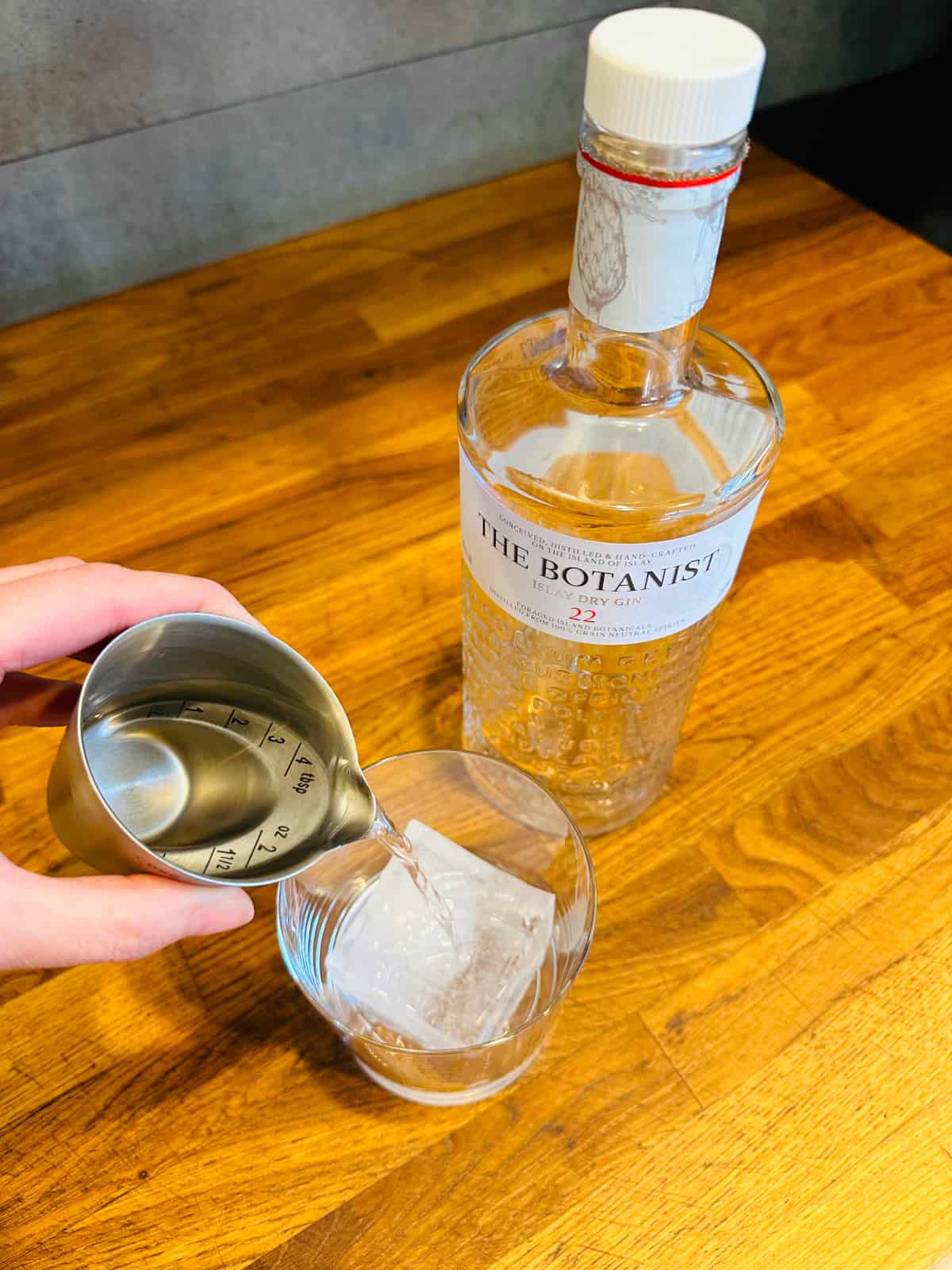Clear liquid being poured from a steel measuring jigger into a rocks glass with a large ice cube next to a bottle of gin.