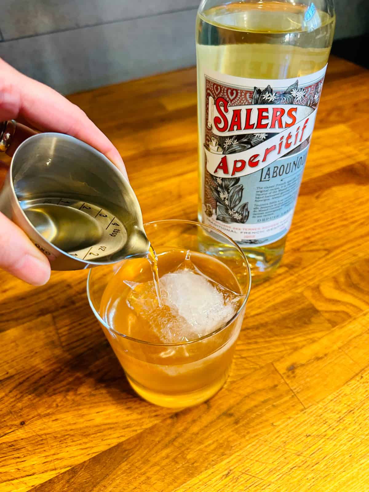 Pale golden liquid being poured from a steel measuring jigger into a rocks glass with a large ice cube next to a bottle of sales.