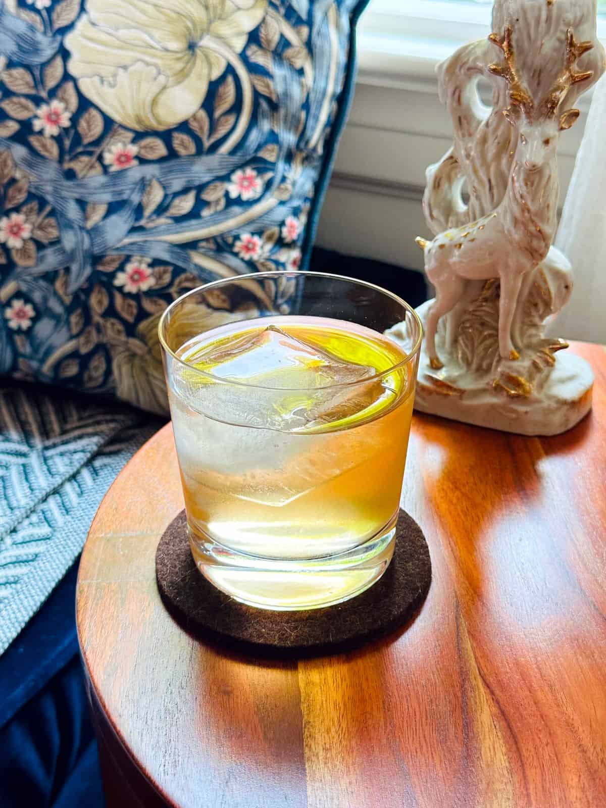 Umeshu Negroni in a rocks glass on a round wooden table next to a porcelain white and gold lamp in the shape of a stag with a blue and white flowered pillow in the background.