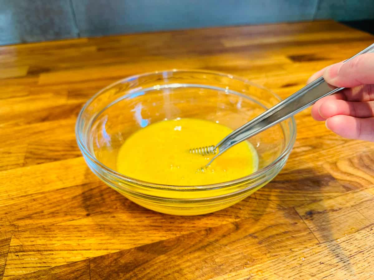 Beaten eggs being beaten with a small steel whisk in a glass bowl.
