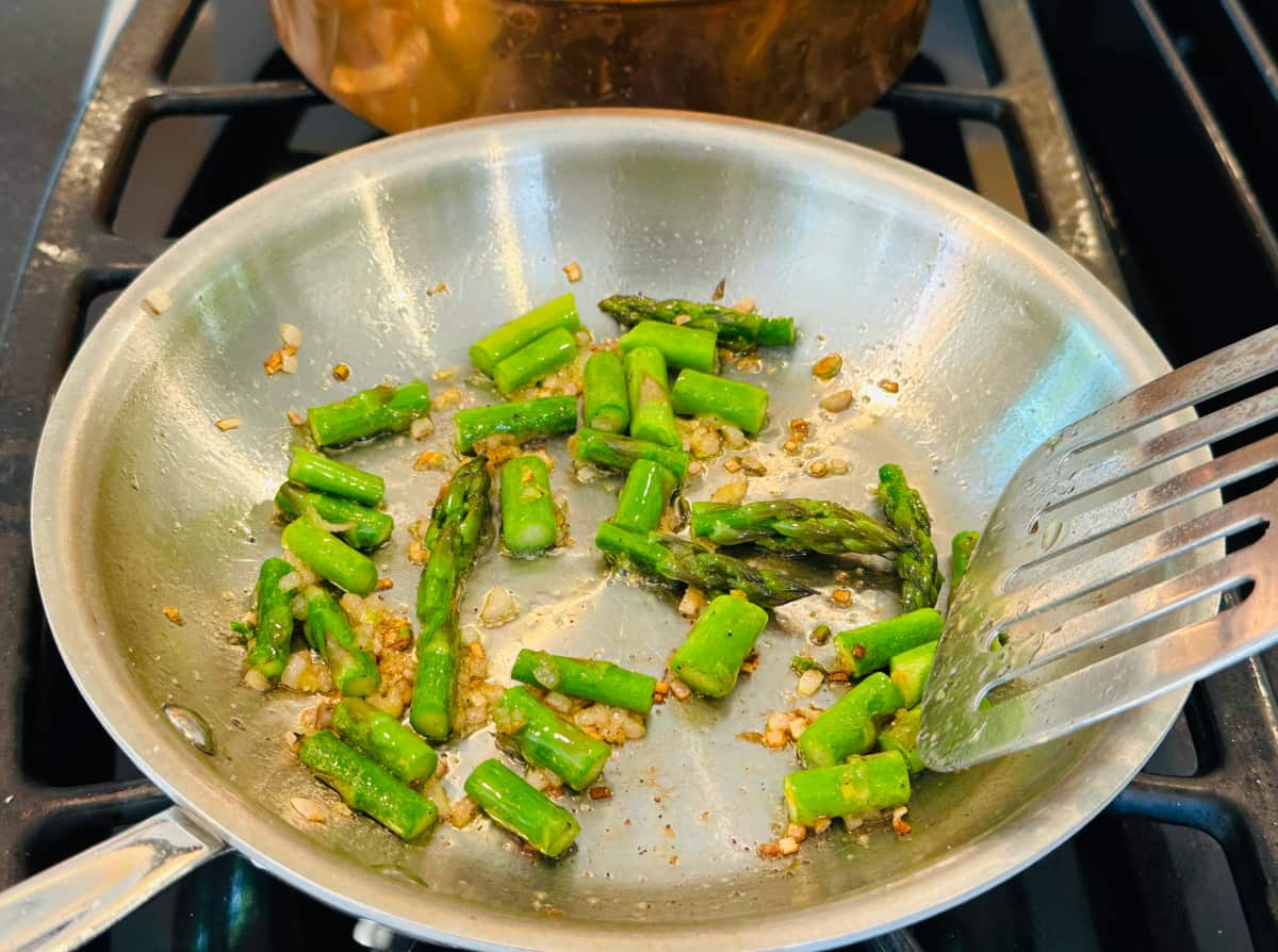 Pieces of bright green asparagus and flecks of browned shallots cooking in a small steel skillet.