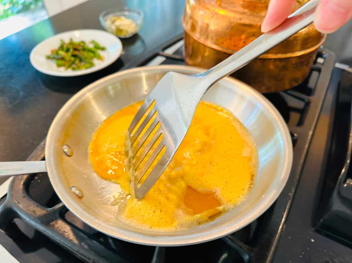 Beaten eggs being stirred with a steel spatula in a small steel skillet.