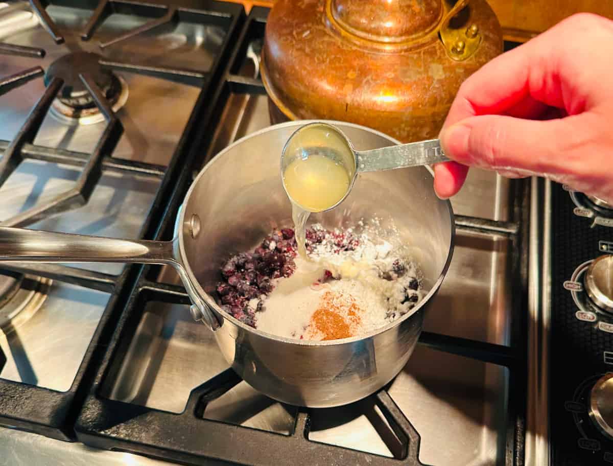 Lemon juice being poured from a measuring spoon into a small steel saucepan with blueberries, sugar, and cinnamon.