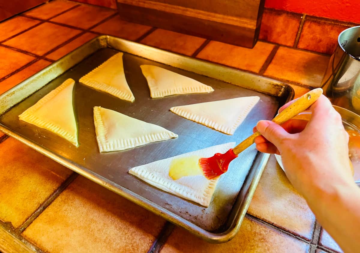 Unbaked turnovers on a metal baking sheet being slicked in egg wash with a red silicone brush.