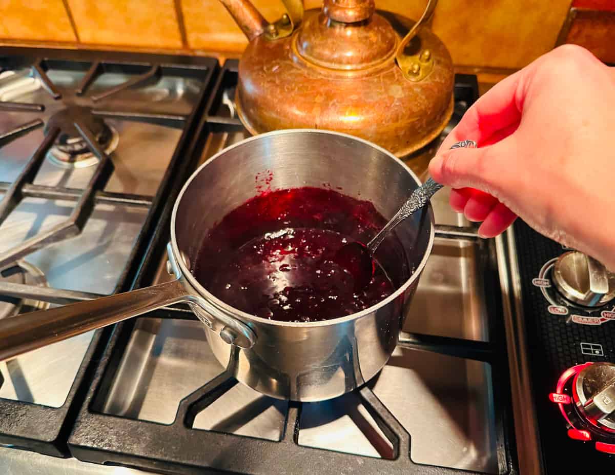 Blueberry filling being simmer and stirred with a spoon in a small steel saucepan.