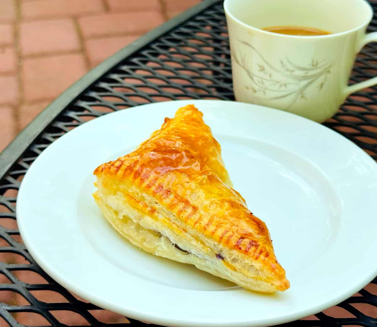 Blueberry turnover on a white plate with a cup of coffee alongside sitting on an black metal outdoor table.