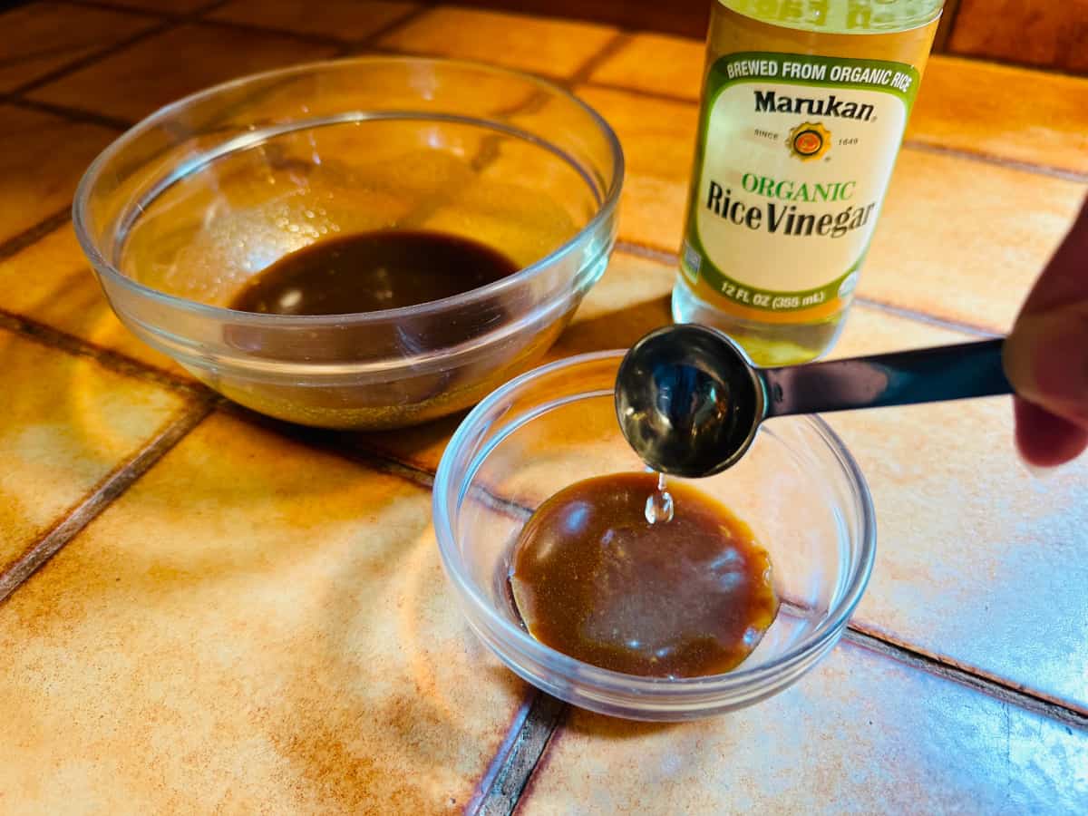 Pale yellow liquid being poured from a measuring spoon into a small bowl of dark brown glaze next to a bottle of rice vinegar.