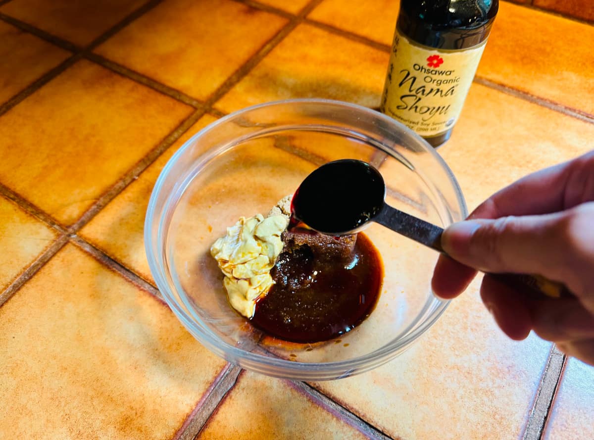 Dark brown liquid being poured from a measuring spoon into a small glass bowl with brown sugar and mustard next to a bottle of shoyu.