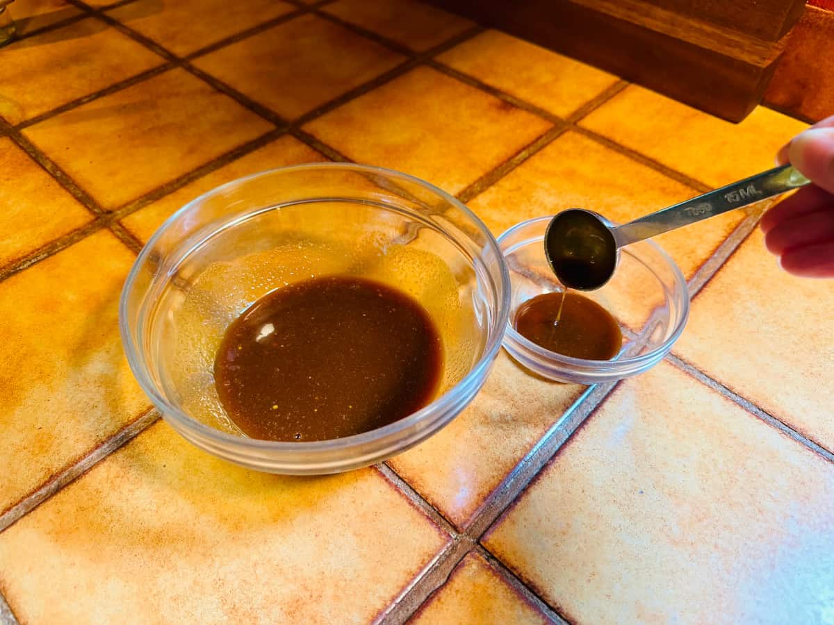 Brown liquid being poured from a measuring spoon into a small glass bowl sitting next to a medium glass bowl containing the same brown liquid.