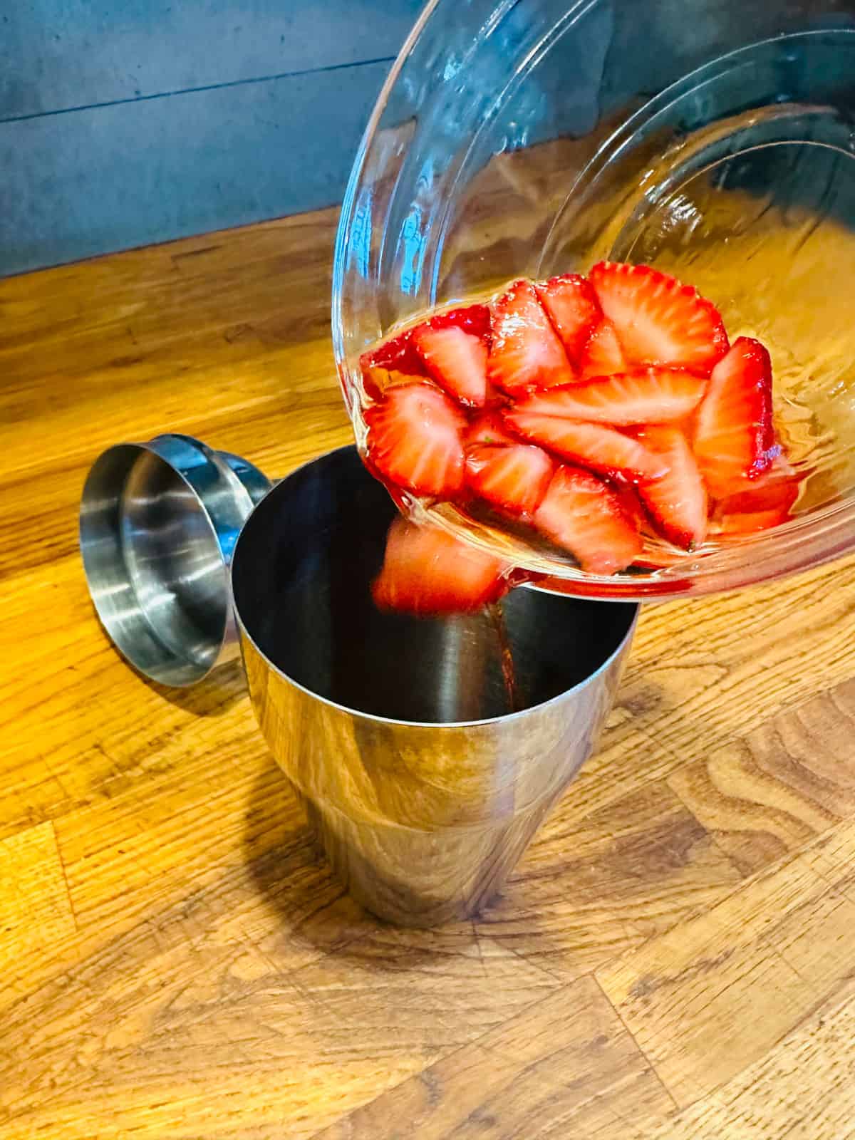 Sliced strawberries in pale liquid being poured from a glass bowl into a metal cocktail shaker.
