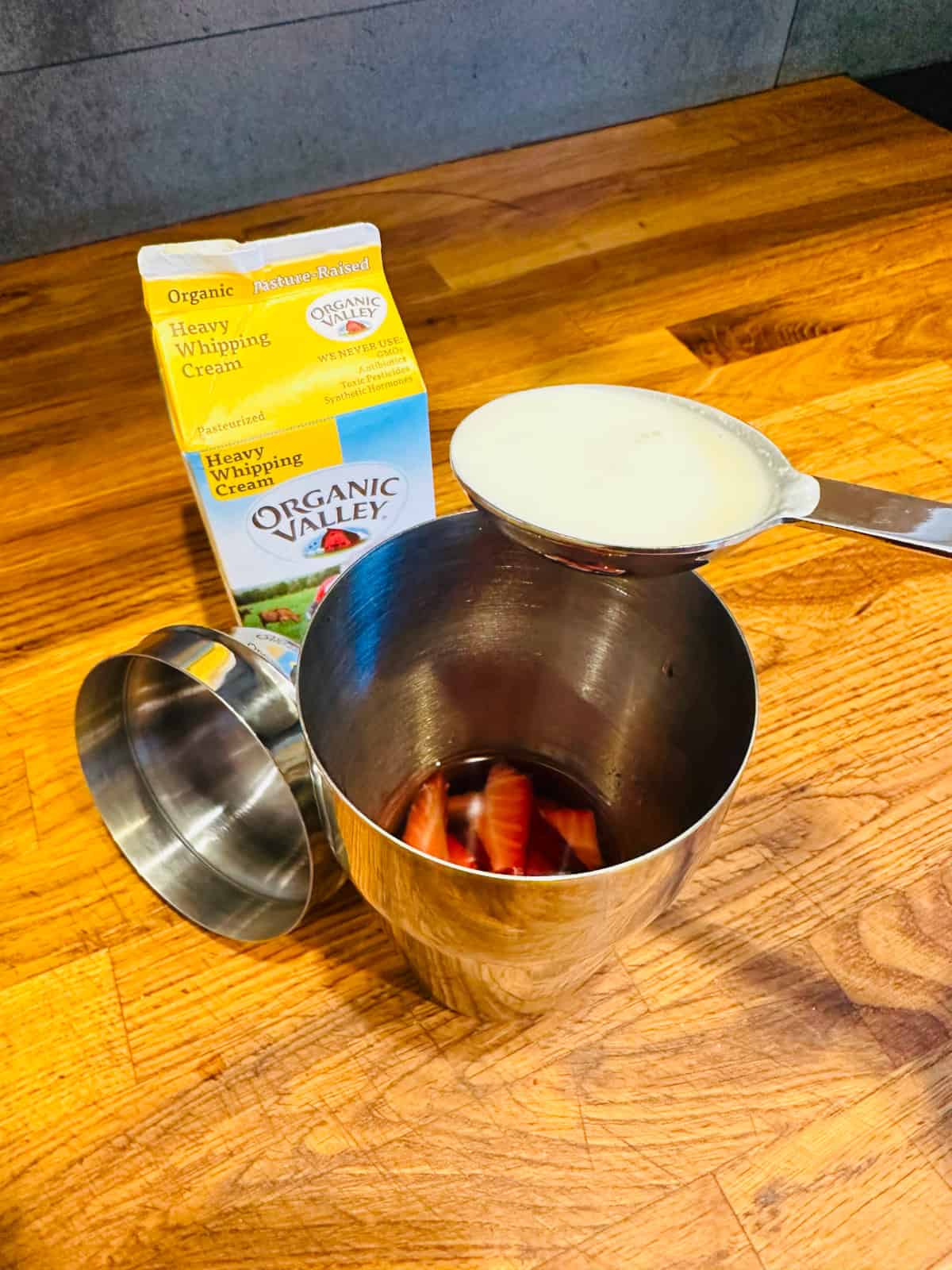A teaspoon of cream being held over a cocktail shaker next to a carton of heavy whipping cream.