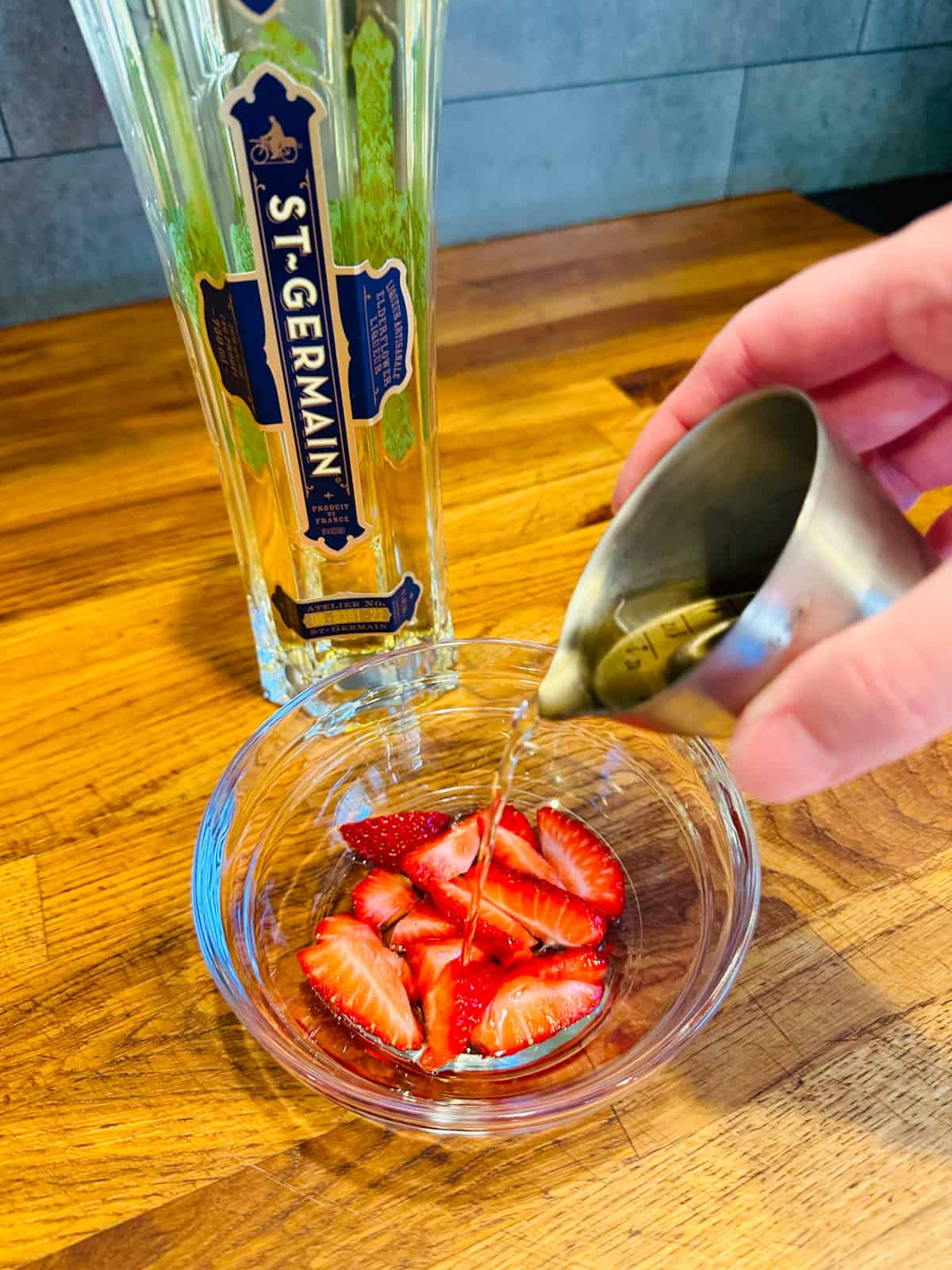 Pale golden liquid being poured from a steel measuring jigger over sliced strawberries in a small glass bowl next to a bottle of St. Germain.