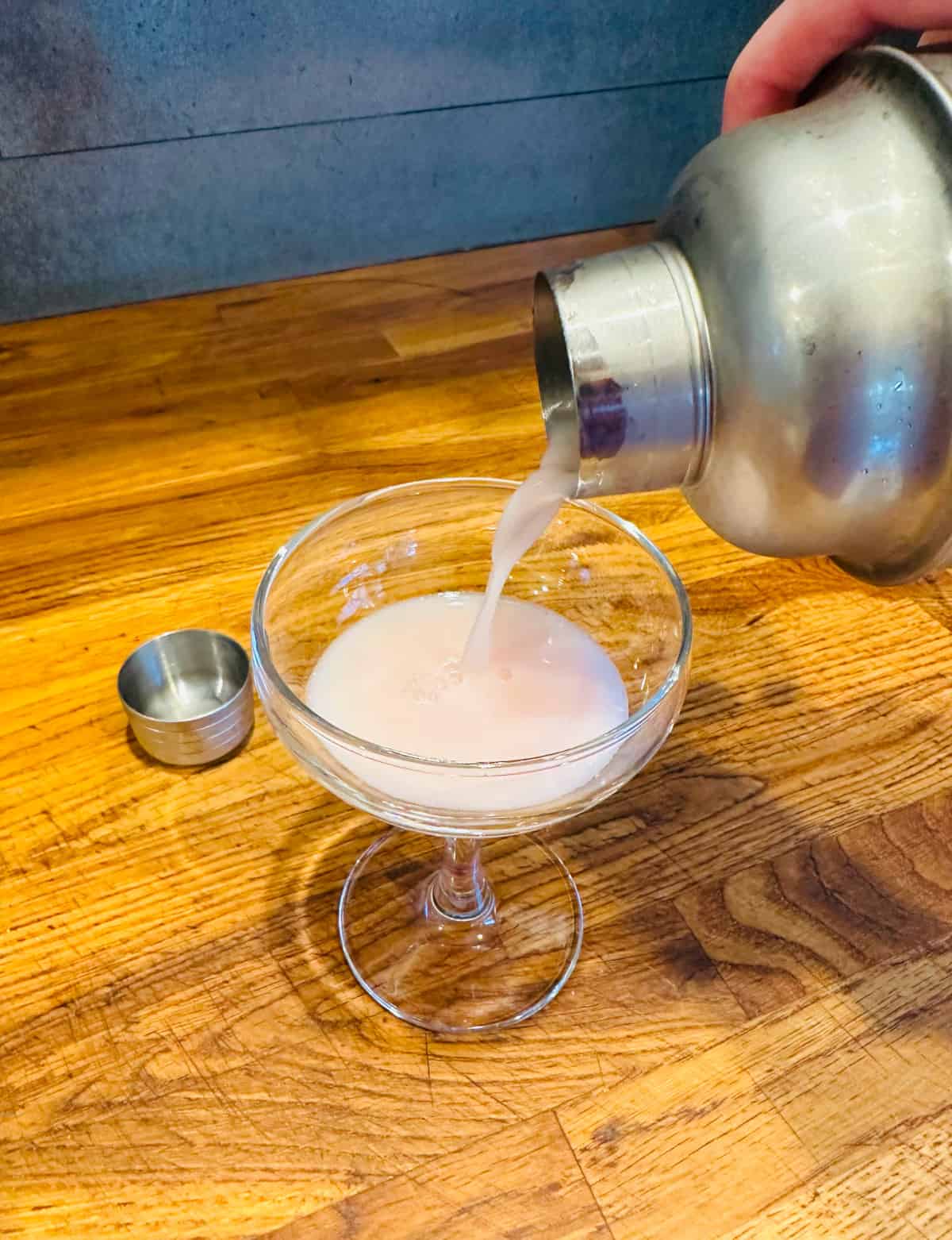 Pale pink liquid being poured from a cocktail shaker into a coupe glass.