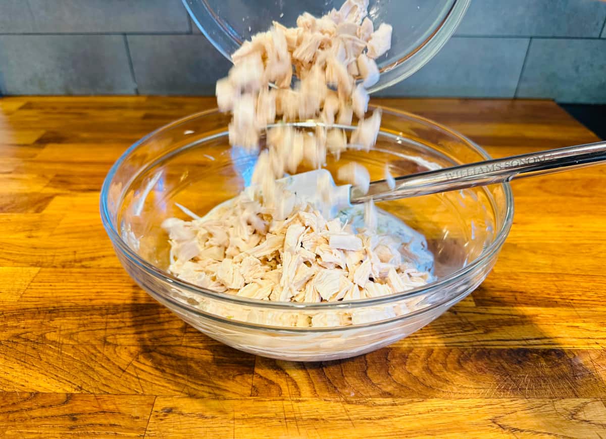 Pieces of cooked chicken being transferred from a glass bowl into a larger glass bowl with mayonnaise mixture and a spatula.