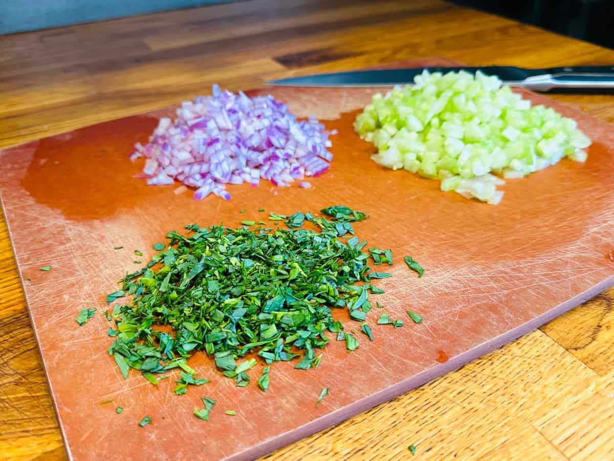 Chopped tarragon, red onion, and celery on a cutting board with a chef's knife.