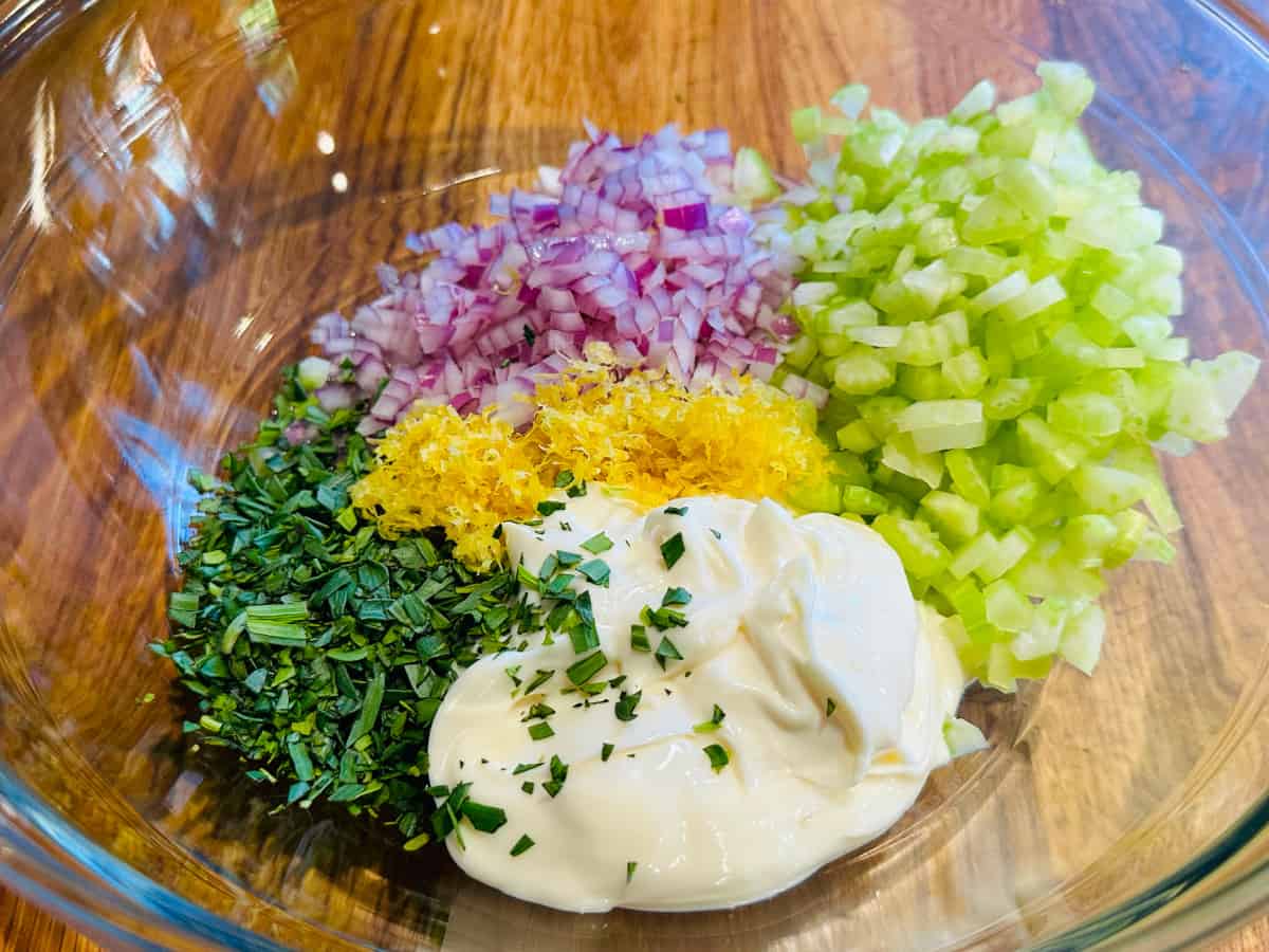 Mounds of chopped tarragon, red onion, and celery along with lemon zest and mayonnaise in a glass bowl.