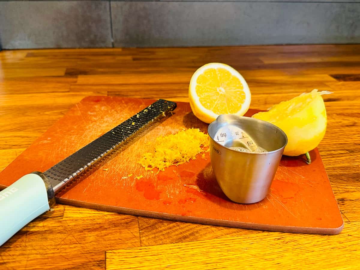 Microplane grater, a small pile of lemon zest, two lemon halves, and a steel measuring jigger sitting on a small cutting board.