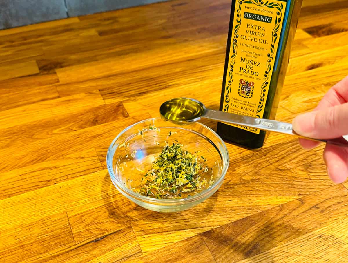 Pale golden liquid in a steel teaspoon being held over a small glass bowl with a mixture of lemon zest, thyme, and garlic next to a bottle of olive oil.
