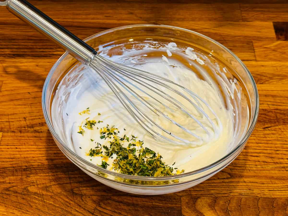 Creamy white goat cheese mixture in a glass bowl with a steel whisk and a sprinkling of lemon zest, thyme, and garlic on top.