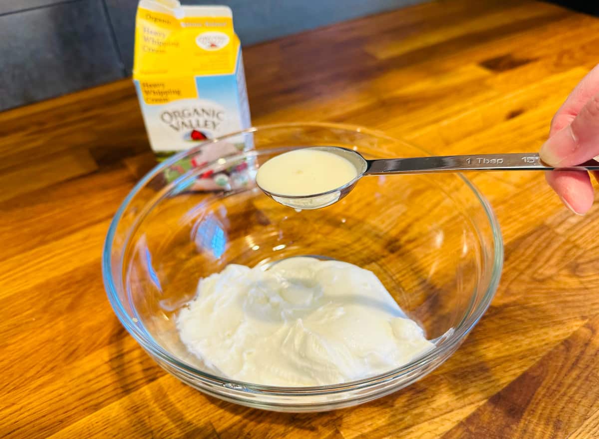 White liquid in a steel tablespoon being held over a bowl of goat cheese in front of a carton of heavy whipping cream.