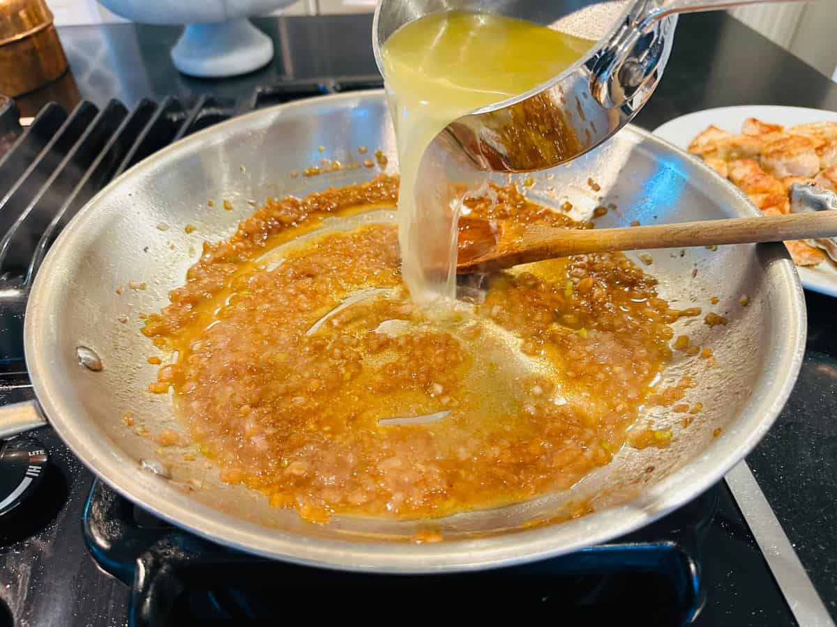 Yellow liquid being poured from a small steel saucepan into sautéed shallots and cognac in a large steel skillet.