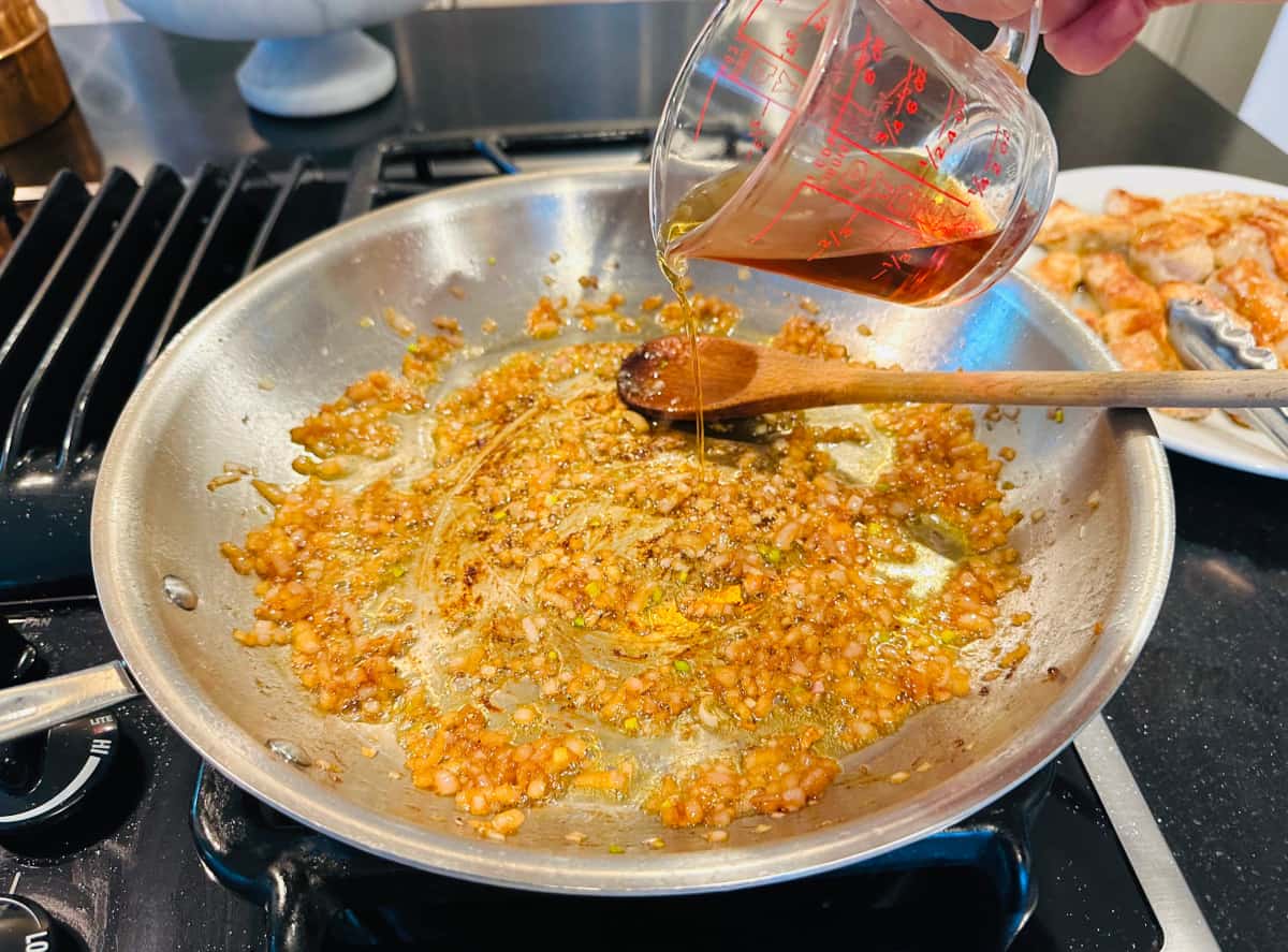 Brown liquid being poured from a glass measuring cup into sautéed shallot in a large steel skillet with a wooden spoon.