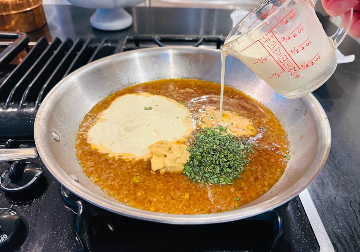 Whipping cream being poured from a glass measuring cup into a large steel skillet containing brown liquid and a small pool of mascarpone, mustard,  and herbs.