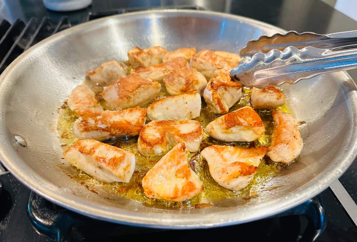 Browned pieces of chicken being turned with metal tongs in a large steel skillet.
