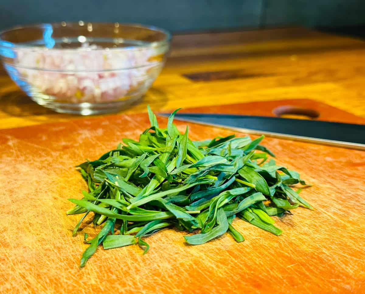 Pile of tarragon leaves in front of a small bowl of chopped shallot and a chef's knife.