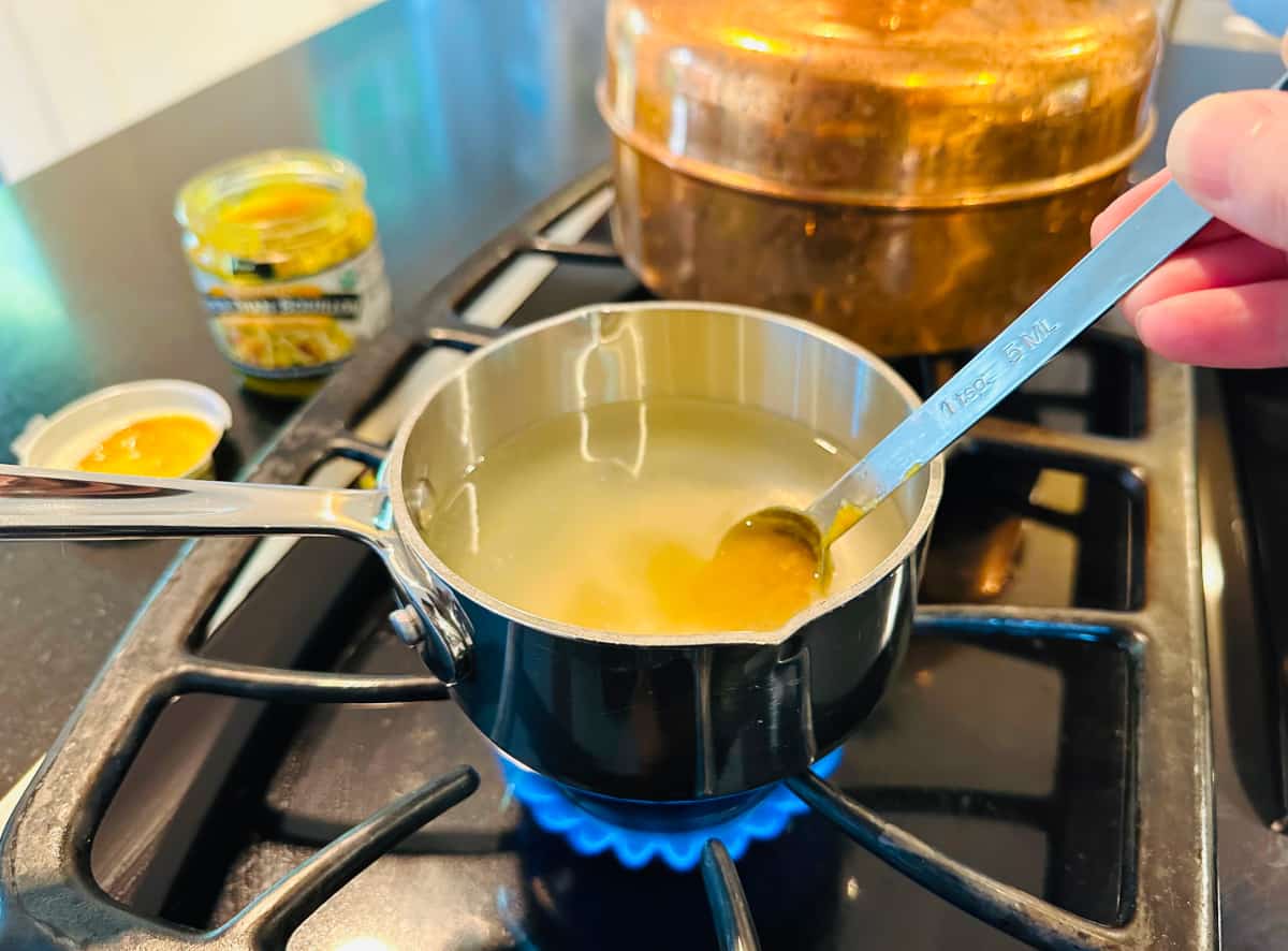 Water and chicken bouillon being stirred with a long measuring spoon in a small steel saucepan on the stove.