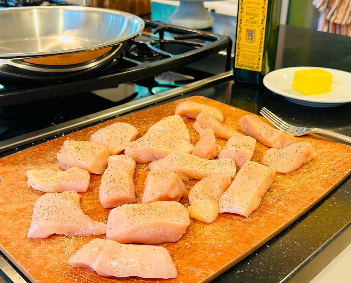 Raw pieces of chicken seasoned with salt and pepper sitting on a cutting board next to a steel skillet on the stove, a bottle of olive oil, and a plate of butter.