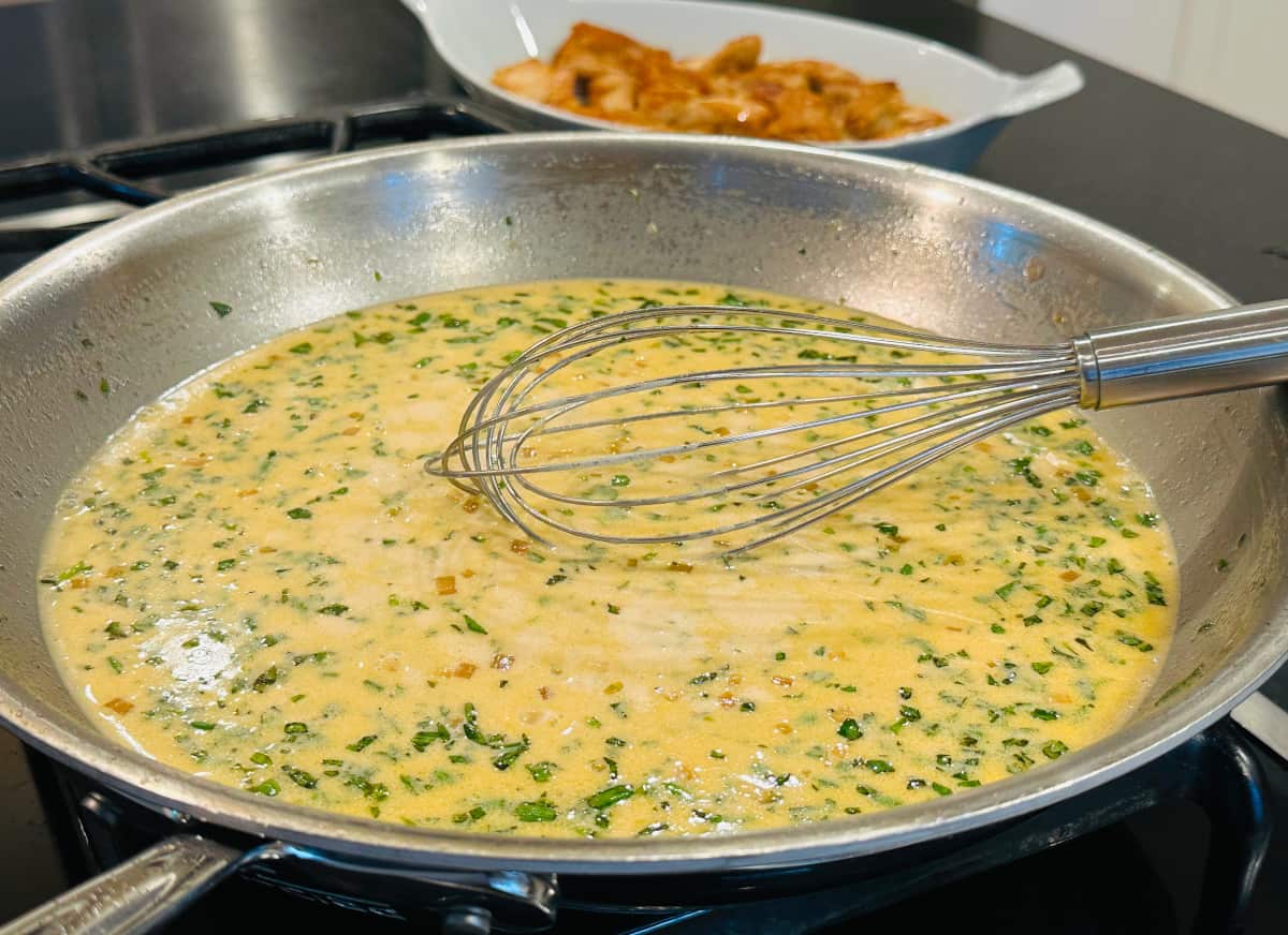 Yellow liquid flecked with green bits of herb simmering in a large steel skillet with a metal whisk.