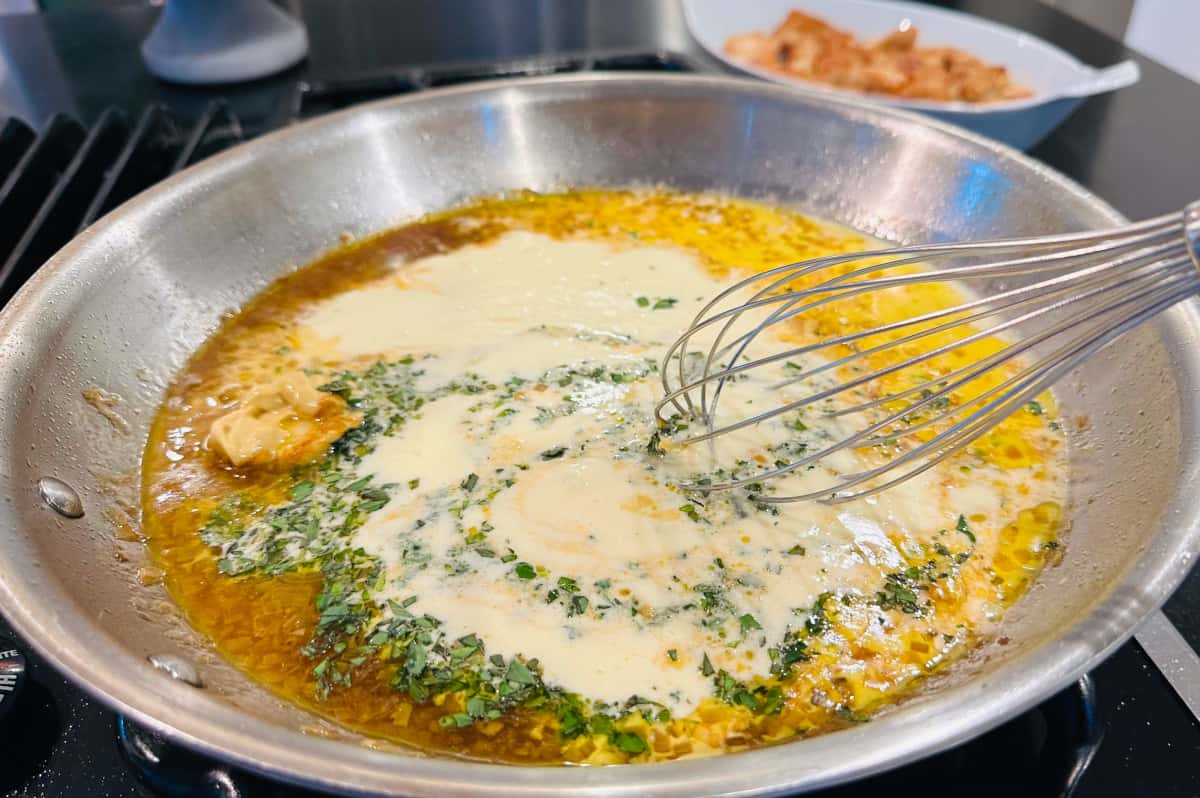 Mascarpone, herbs, mustard, and whipping cream being whisked into broth and cognac in a large steel skillet.
