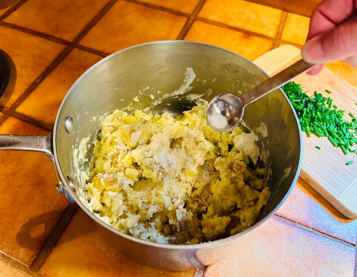 Salt being sprinkled from a teaspoon into a large saucepan of mashed potatoes next to chopped chives on a cutting board.