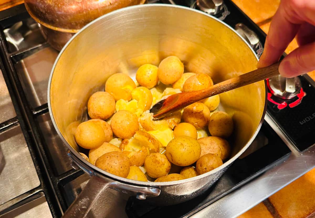 Small yellow potatoes and butter being mashed with a wooden spoon in a large steel saucepan on the stove.