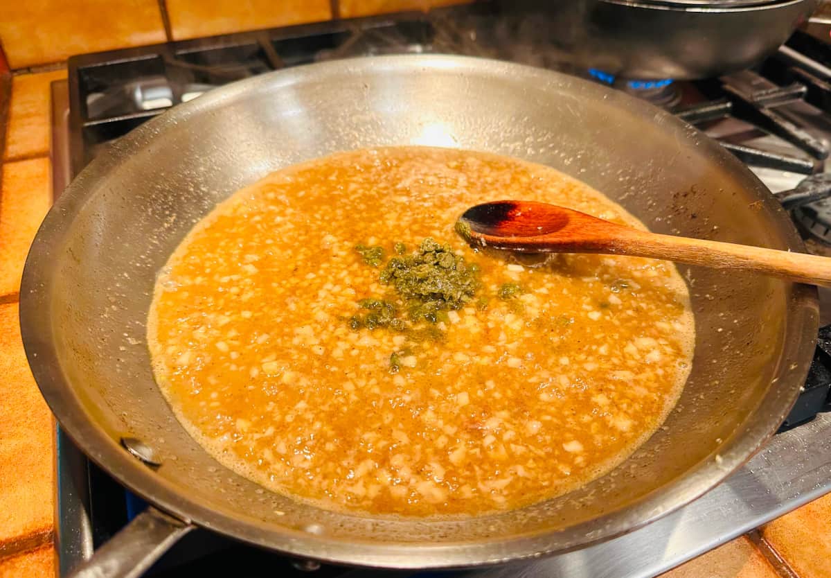 Chopped capers in the center of a large steel skillet of golden brown sauce with a wooden spoon.