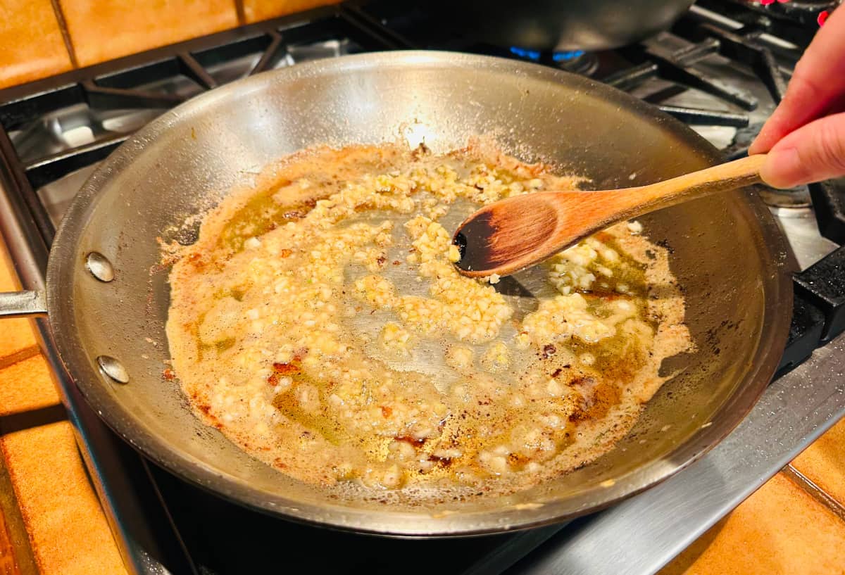 Minced garlic being stirred with a wooden spoon while frying in butter and oil in a large steel skillet.