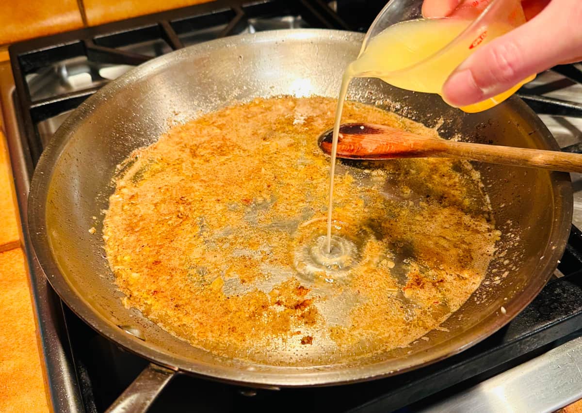 Lemon juice being poured from a plastic measuring jigger into a large steel skillet with browned butter, oil, and white wine.