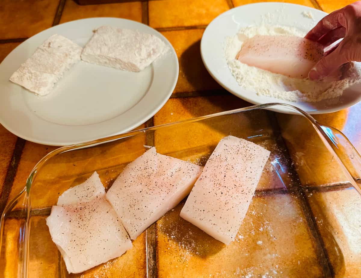 Large chunks of halibut being dredged in a shallow white bowl of flour and then placed on a clean white plate.