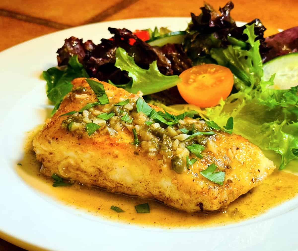 Halibut piccata in front of a green salad on a white plate.