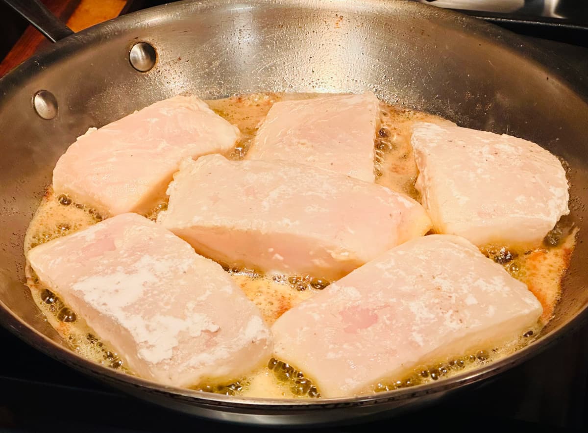 Thick chunks of halibut frying in butter and oil in a large steel skillet.