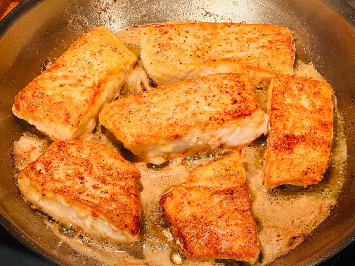 Browned chunks of halibut frying in butter and oil in a large steel skillet.