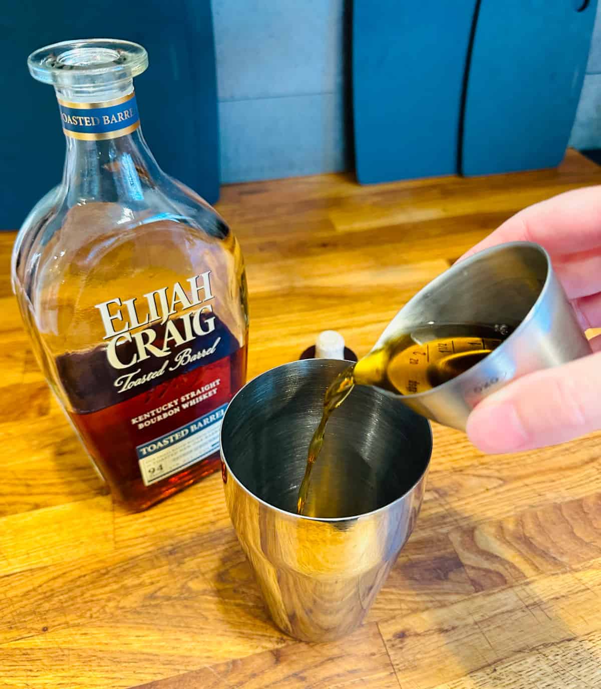 Brown liquid being poured from a steel measuring jigger into a cocktail shaker next to a bottle of bourbon.
