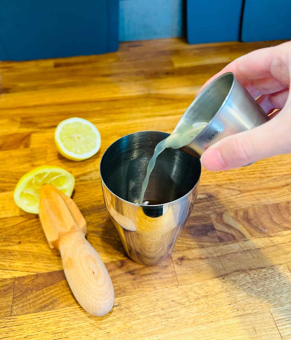 Pale yellow liquid being poured from a steel measuring jigger into a cocktail shaker next to a wooden citrus reamer and two halves of lemon.