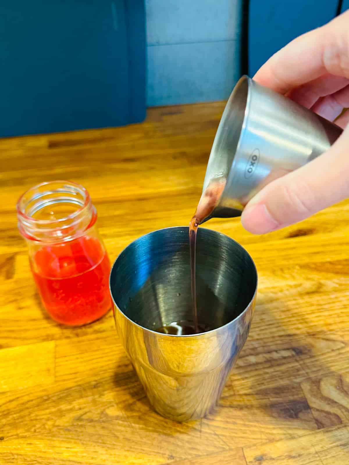 Pink liquid being poured from a steel measuring jigger into a cocktail shaker next to a small jar of pink liquid.