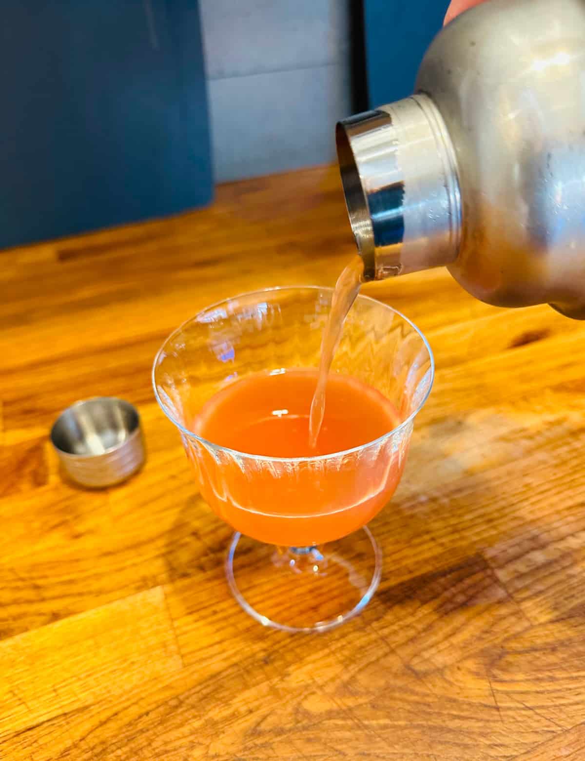 Pinkish orange liquid being poured from a cocktail shaker into a coupe glass.