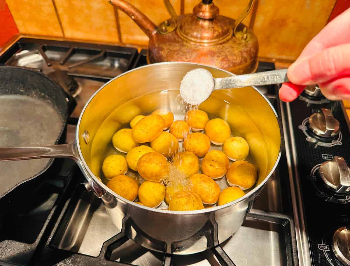 Salt being sprinkled from a teaspoon into a large steel saucepan of potatoes covered with water.