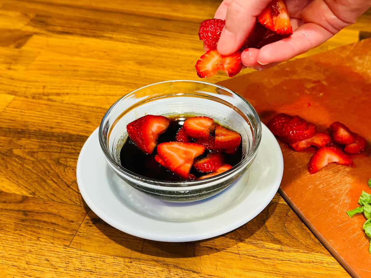 Sliced strawberries being dropped into dark liquid in a small glass bowl sitting on a small white plate.