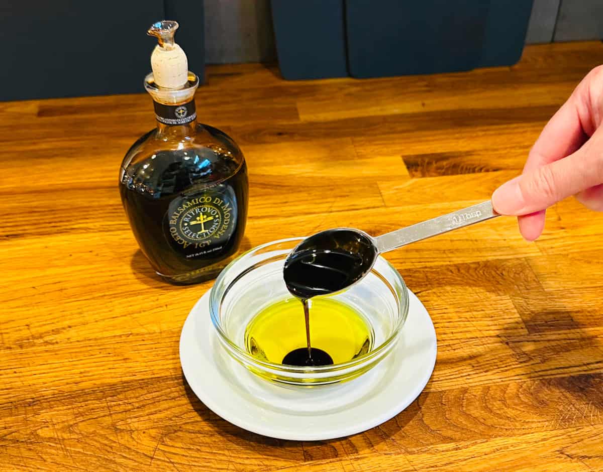 Balsamic vinegar being poured from a steel measuring spoon into olive oil in a small glass bowl sitting on a white plate next to a bottle of balsamic vinegar.